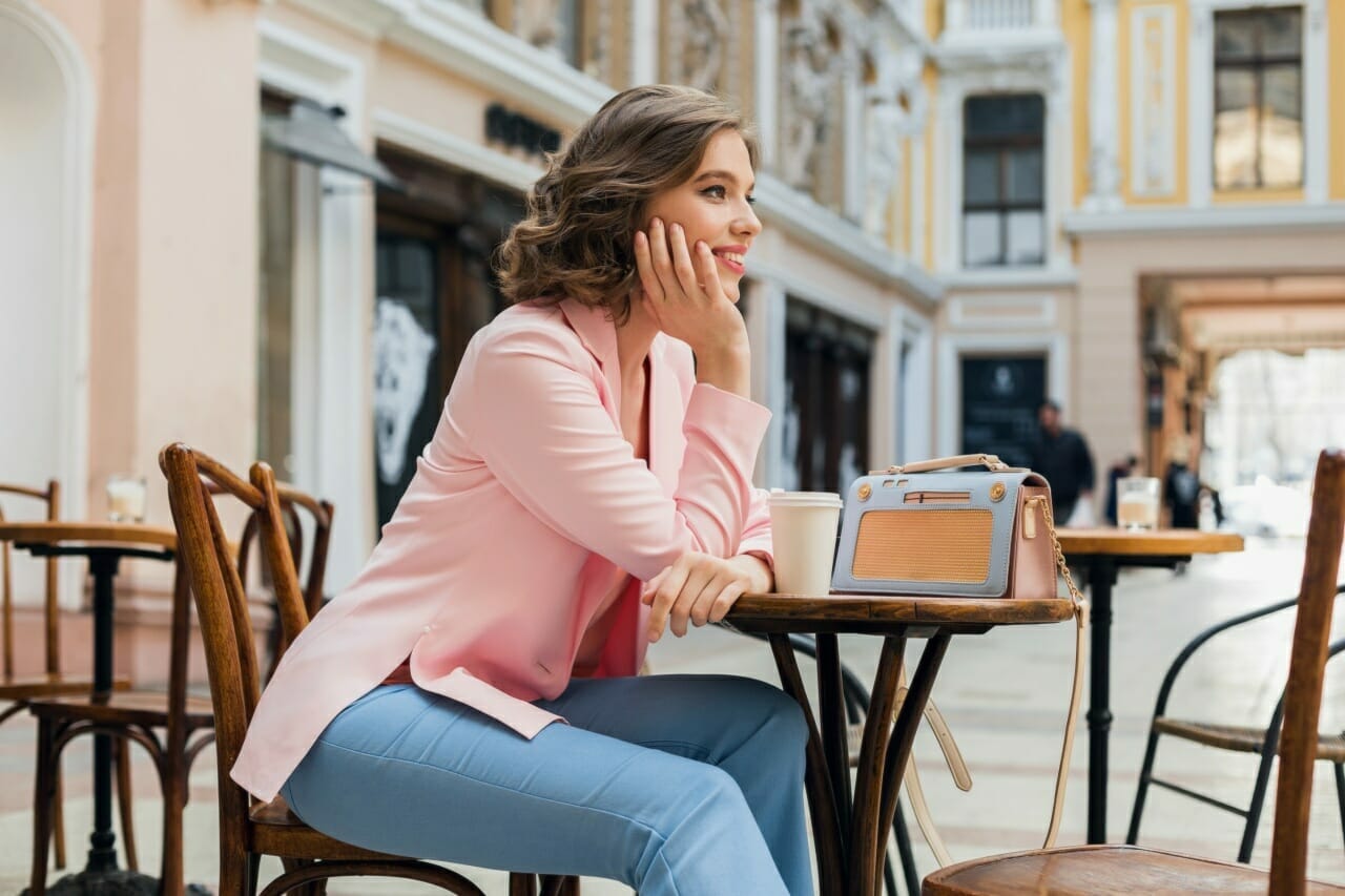portrait-of-stylish-smiling-lady-sitting-at-table-drinking-coffee-in-pink-jacket-summer-style-trend-blue-handbag-accessories-street-style-women-fashion